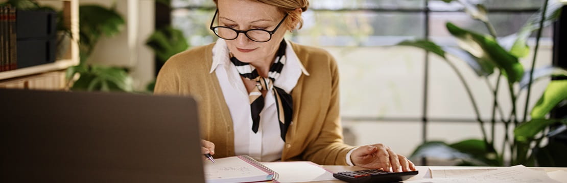 A person sits at a desk in a bright office space with large windows and green plants, working on a laptop while writing in a notebook and using a calculator surrounded by papers.